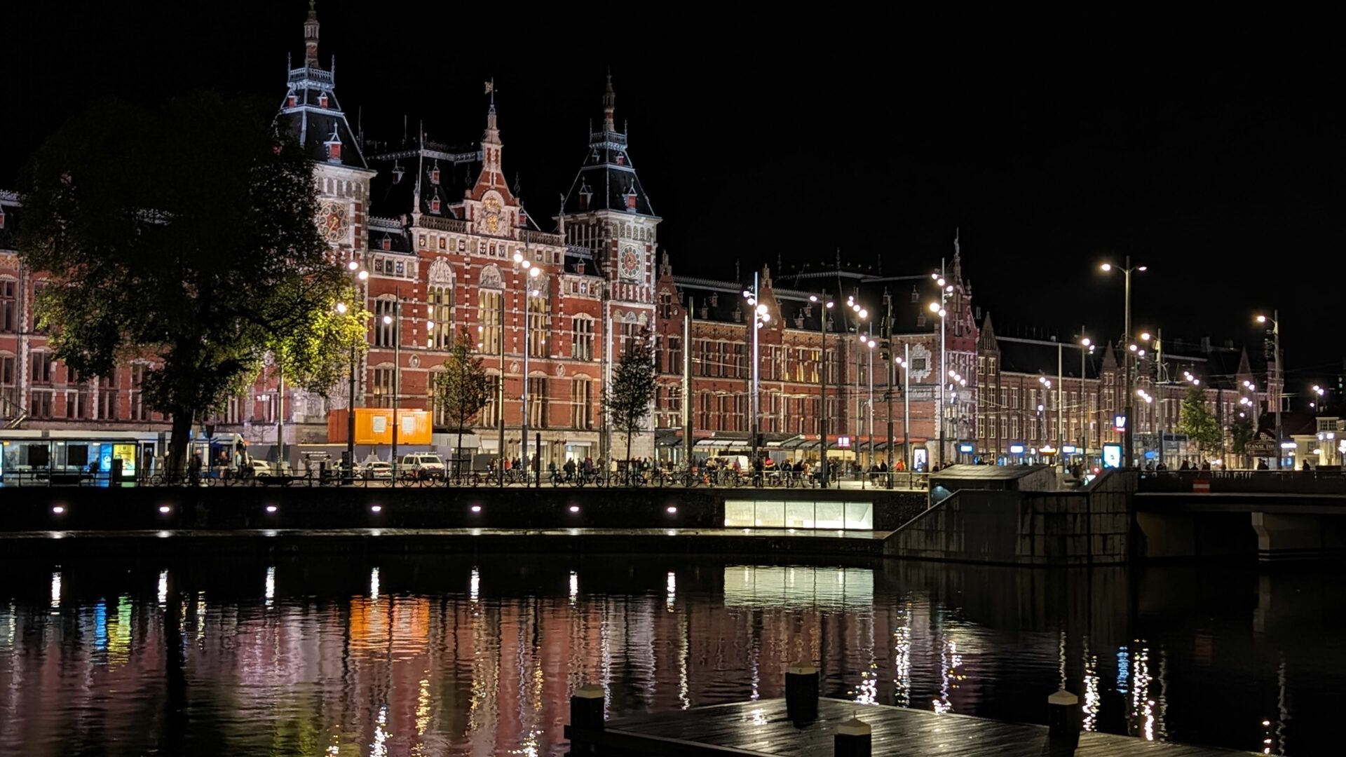 Eine nächtliche Aufnahme des Bahnhofs Amsterdam Centraal, beleuchtet in warmem Licht. Das Gebäude im gotischen und Renaissance-Stil spiegelt sich im Wasser eines Kanals im Vordergrund. Im Hintergrund ist der dunkle Himmel zu sehen.