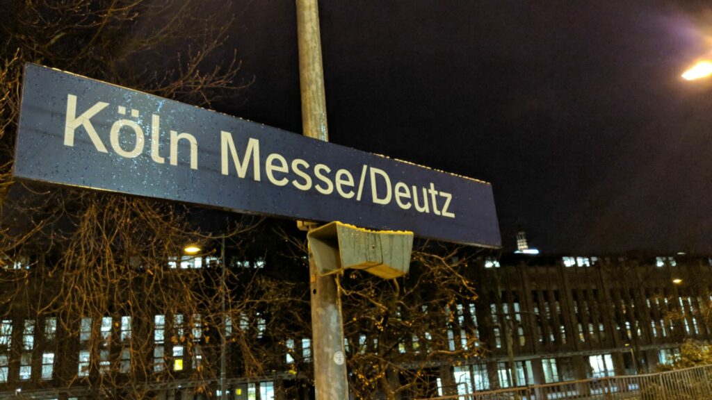 Ein Straßenschild mit der Aufschrift "Köln Messe/Deutz" in weißen Buchstaben auf blauem Grund. Das Schild ist an einem Pfosten befestigt und nachts von unten beleuchtet. Im Hintergrund sind verschwommen die beleuchteten Fenster eines Gebäudes und die kahlen Äste eines Baumes zu sehen.